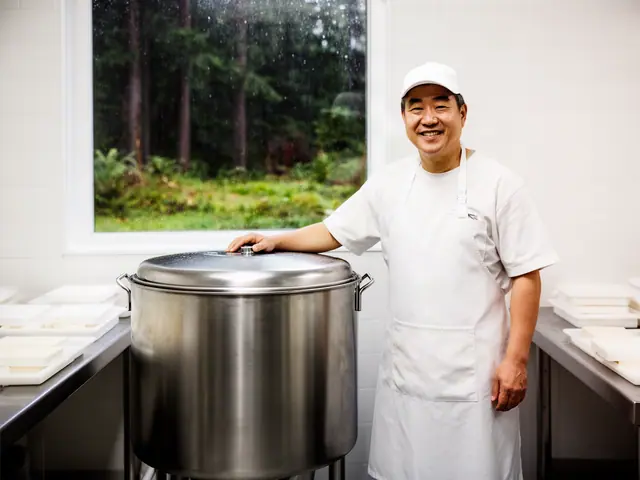 Smiling man in a white cap and apron standing beside a large stainless steel pot in a bright white kitchen