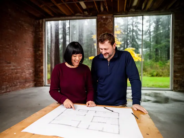 Two business partners smiling over a floor plan on a wooden table in a bare brick building, rainy forest window behind